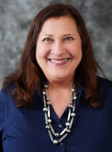 Image is a smiling woman with medium-length brown hair. She is wearing a blue button-up shirt and a silver beaded necklace.